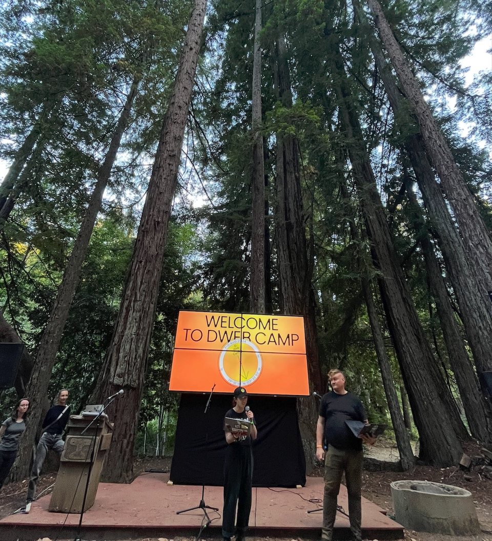 Mai and Ian at DWeb Camp 2024 with a screen that says Welcome to DWeb Camp behind them, and towering redwoods