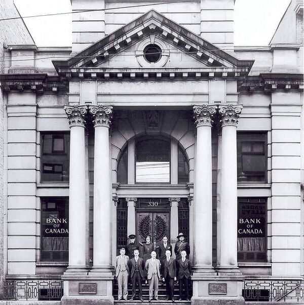 Black and white photo of a close cropped front of the Permanent. Windows have stencils that read Bank of Canada. There are 9 people in business suits standing on the steps.